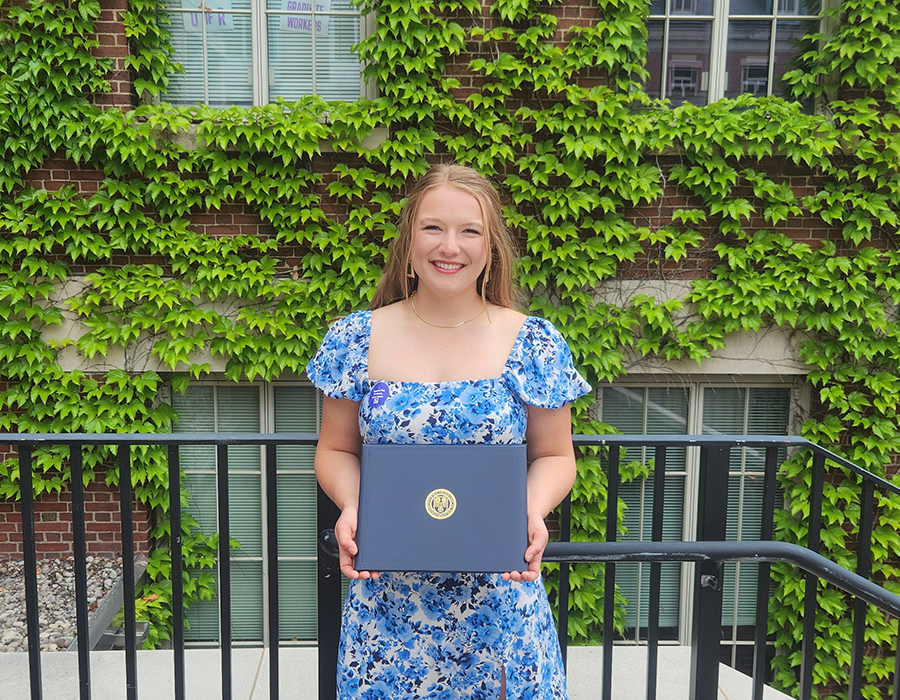 Sydney in front of a brick building smiling at the camera wearing a floral dress and holding a certificate.
