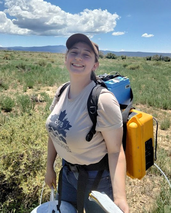 Katie smiling at the camera while walking through a field wearing a backpack.