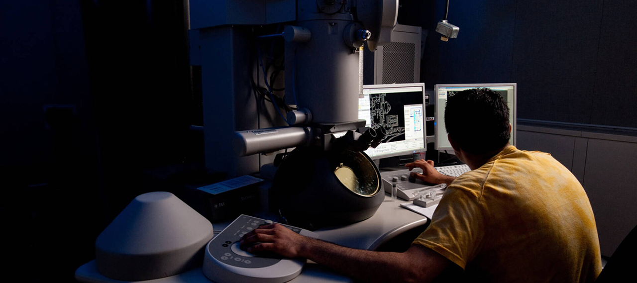A person in a yellow shirt seen from behind works with a transmission electron microscope in a darkened room.