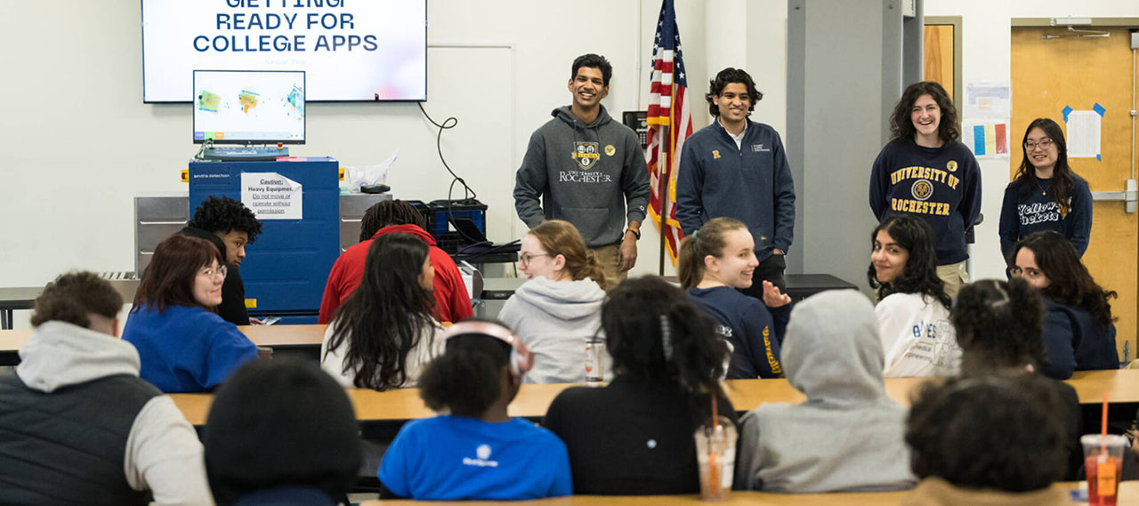 Student educational consultants from Project Level the Field wear University of Rochester gear and stand in front of a room full of high school students with a screen near them that reads.