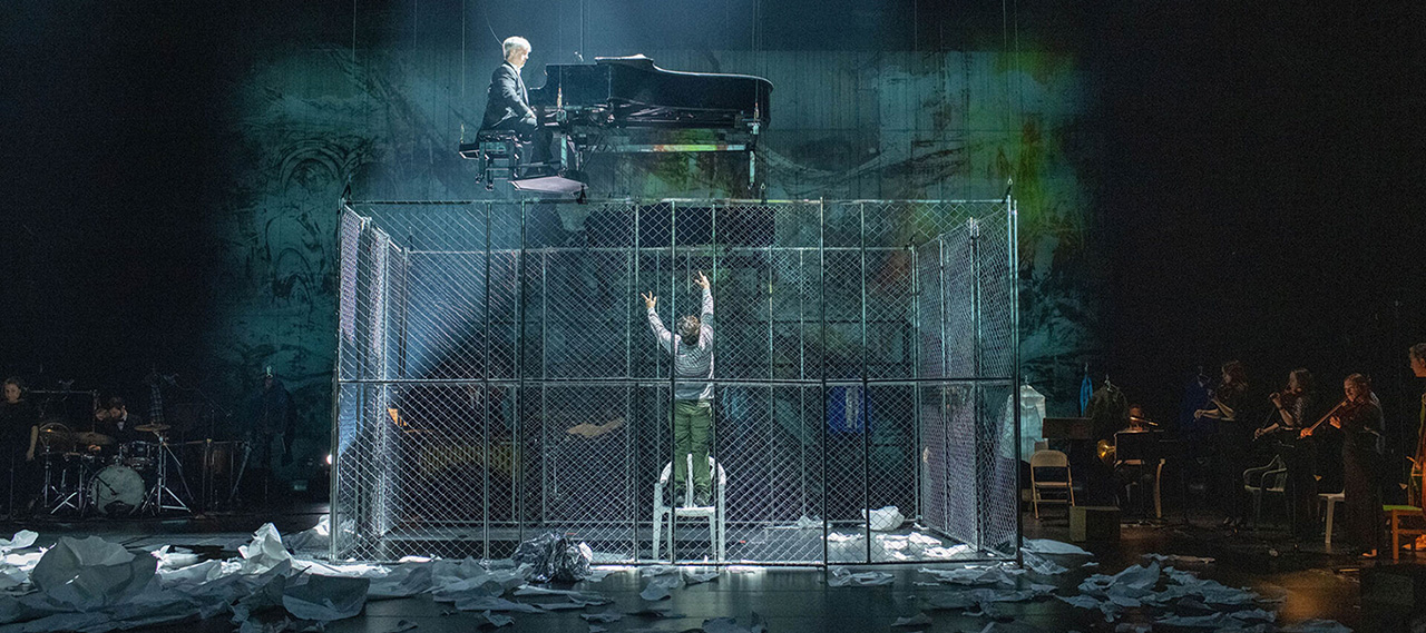 Man in cage reaches up toward piano player in scene from Paper Pianos.