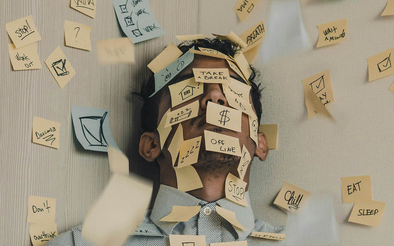 Man's face covered in post-its with the words of common stressors and solutions on them, including money, time, don't overthink, and more.