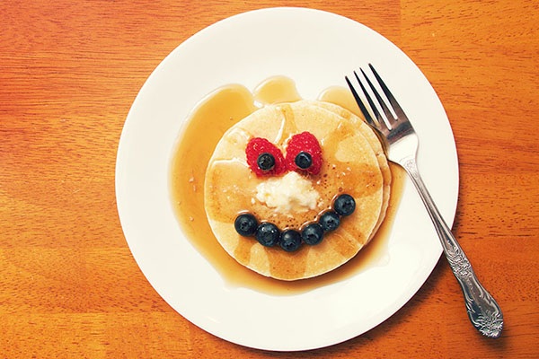 a plate of pancakes and maple syrup making a funny face with fruit.