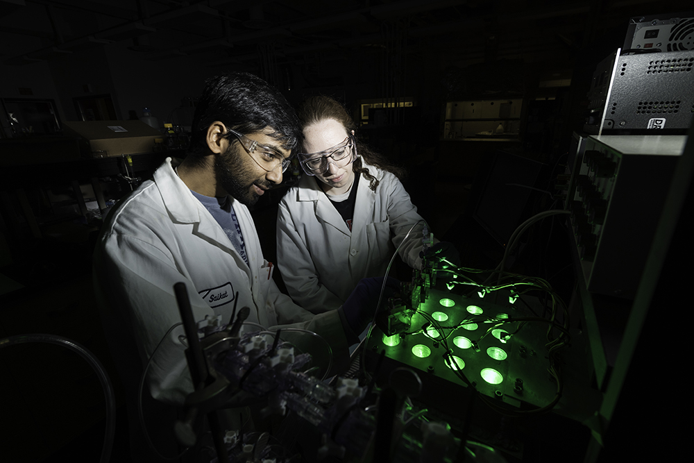Two students working in a dark lab setting with bright green elements.
