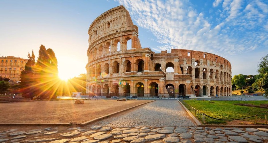 An exterior view of some ancient ruins with the sun setting behind it.