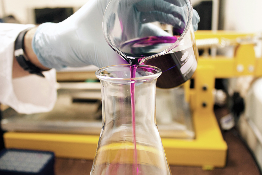 A hand pouring purple liquid into a clear glass beaker.