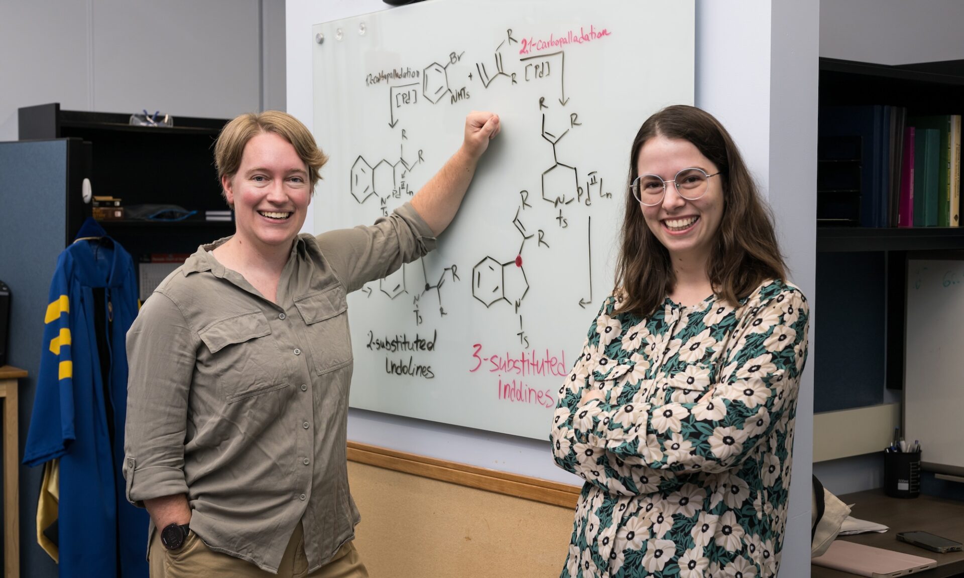 Professor Paradine and graduate student Rodina smiling and facing camera, standing against whiteboard showing chemical formulas.