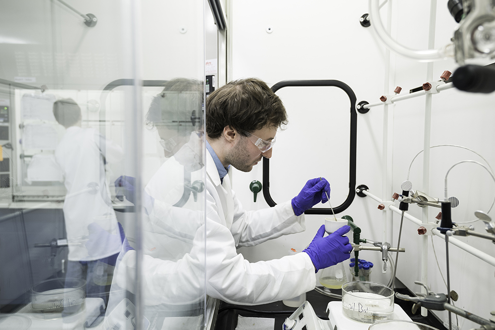 A student wearing a lab coat, gloves, and eye protection is mixing compounds in a lab.