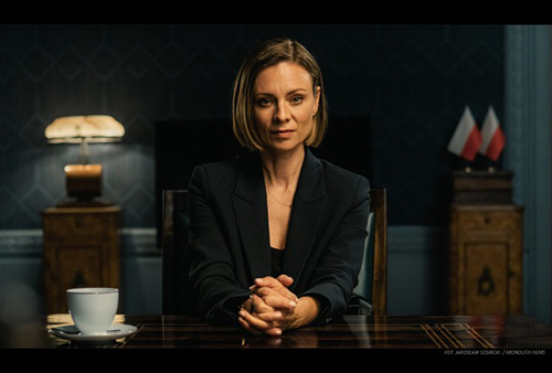 A still image from the film of a woman sitting at a table with a tea cup staring into the camera.