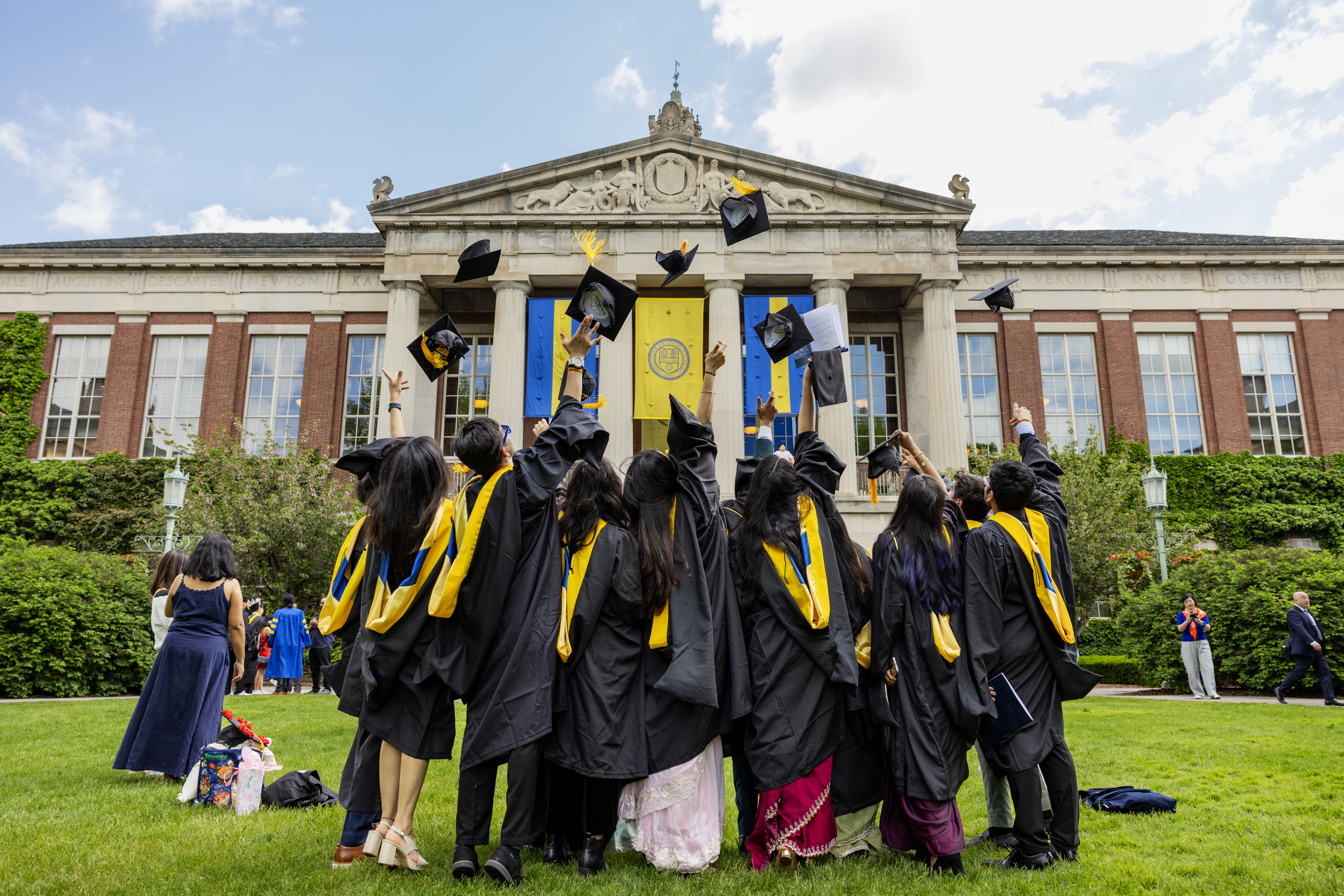 University students in graduation robes, throwing their caps in the air, in front of Rush Rhees Library