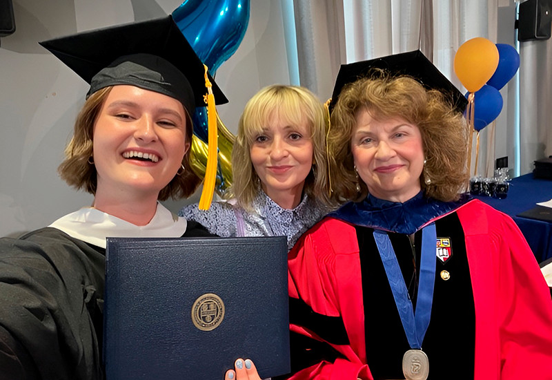 A graduate in cap and gown holds their diploma takes a selfie with two other people.