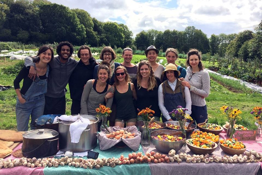 A group of people pose for the camera while standing behind a table laden with food in a field.