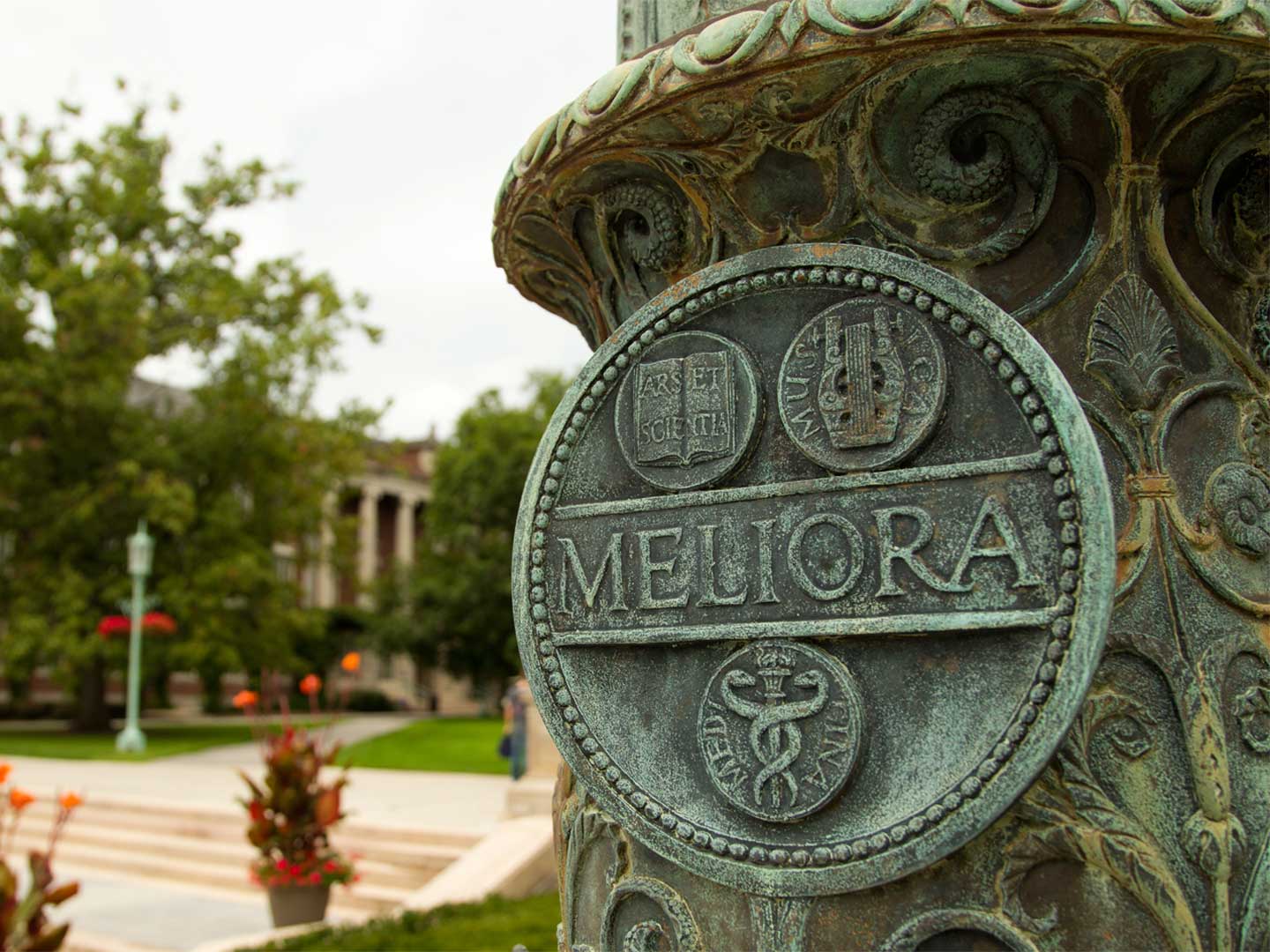 The meliora seal at the base of the flag pole, trees, pillared building, and stairs in the background.