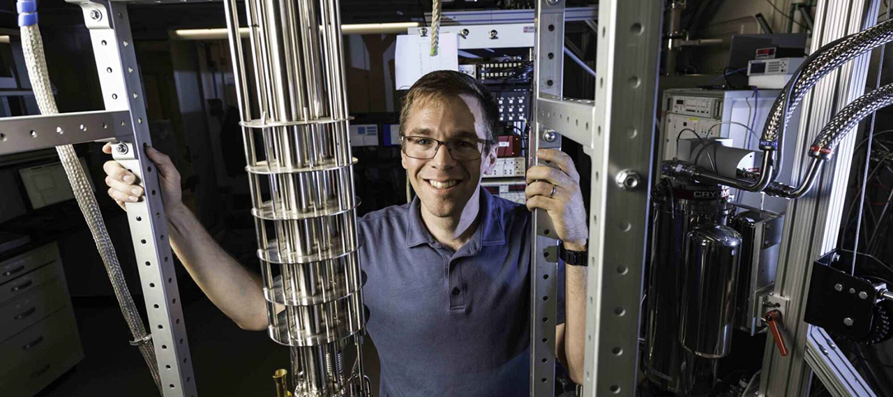 John Nichol standing behind a dilution refrigerator, used to study quantum systems, including nuclear-spin dark states.