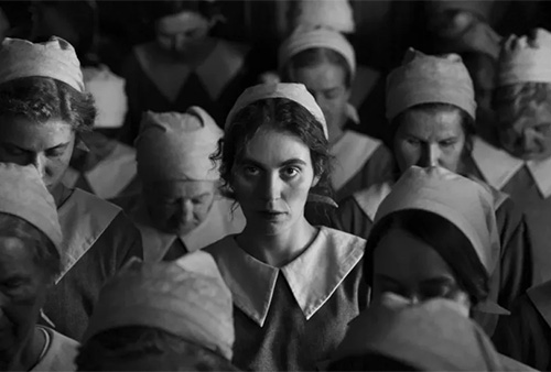 A still image from the film of a woman in staring at the camera surrounded by other women with their heads bowed.